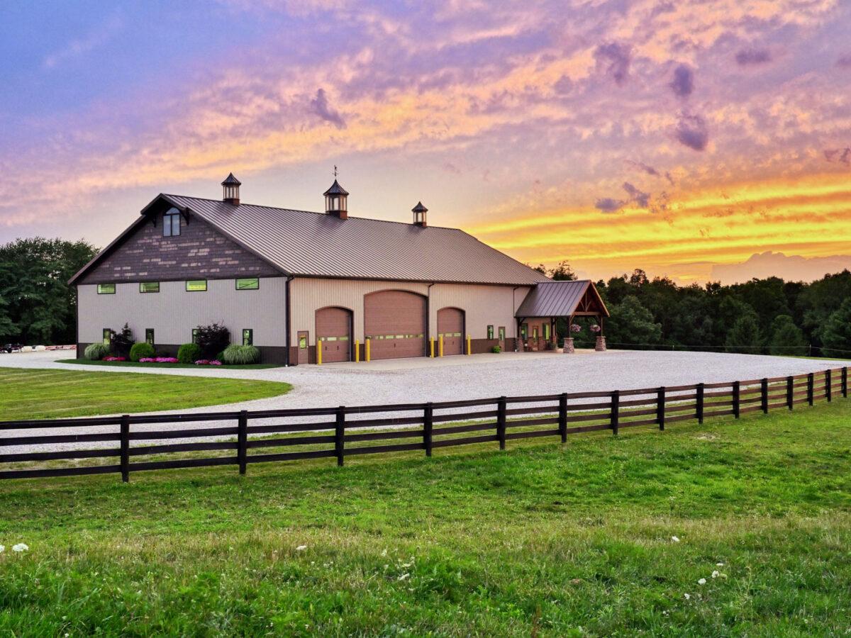 High-end beige agricultural pole barn featuring a brown metal roof, decorative cupolas, and a wraparound porch at sunset.